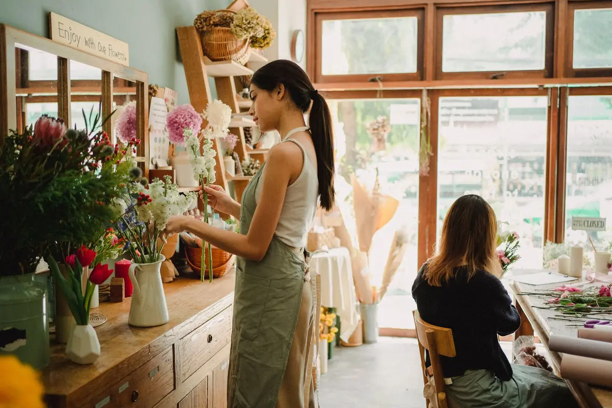 A florist arranging a bouquet in her shop while a customer sits and waits